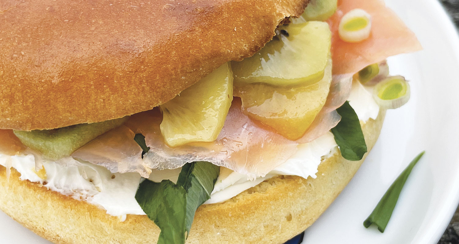 un bagel avec de la truite fumée sur un lit de philadelphia , de l'aillet ciselé, des kiwis jaunes et verts et de l'ail des ours, un bagel de printemps sur une assiette la plage par assiettes et compagnie