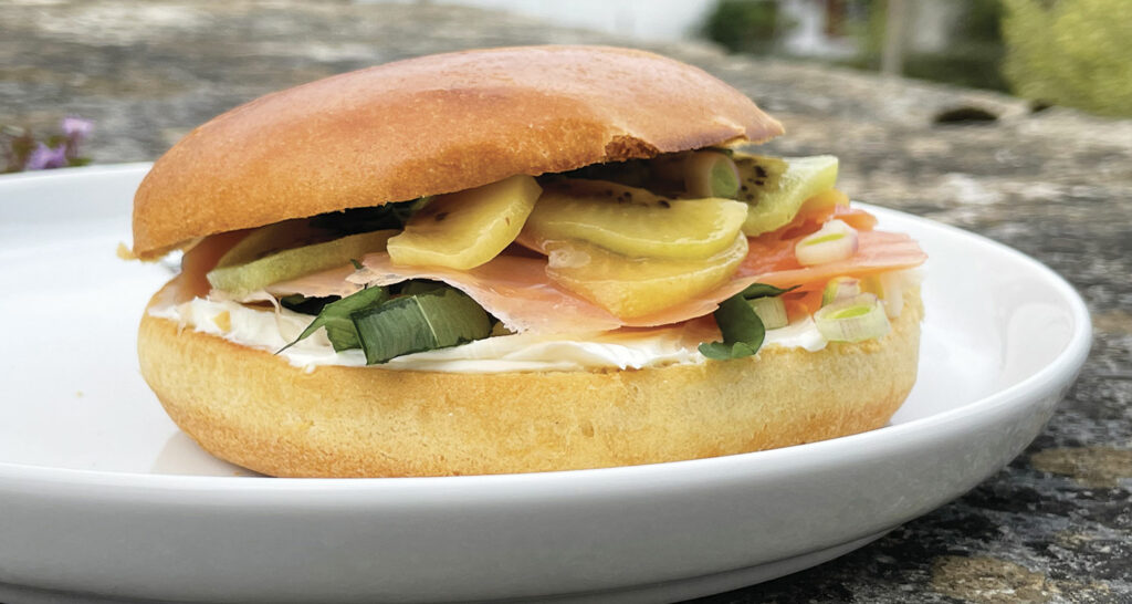 un bagel avec de la truite fumée sur un lit de philadelphia , de l'aillet ciselé, des kiwis jaunes et verts et de l'ail des ours, un bagel de printemps sur une assiette la plage par assiettes et compagnie