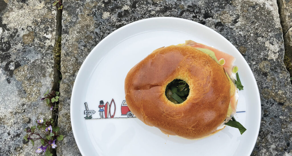 un bagel avec de la truite fumée sur un lit de philadelphia , de l'aillet ciselé, des kiwis jaunes et verts et de l'ail des ours, un bagel de printemps sur une assiette la plage par assiettes et compagnie